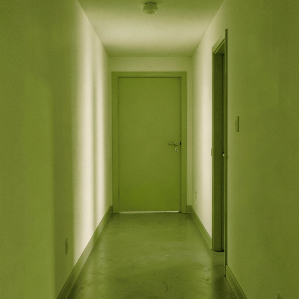 A tall duotone photograph in olive green and cream of a narrow quiet hallway with soft light, calm and plain in mood