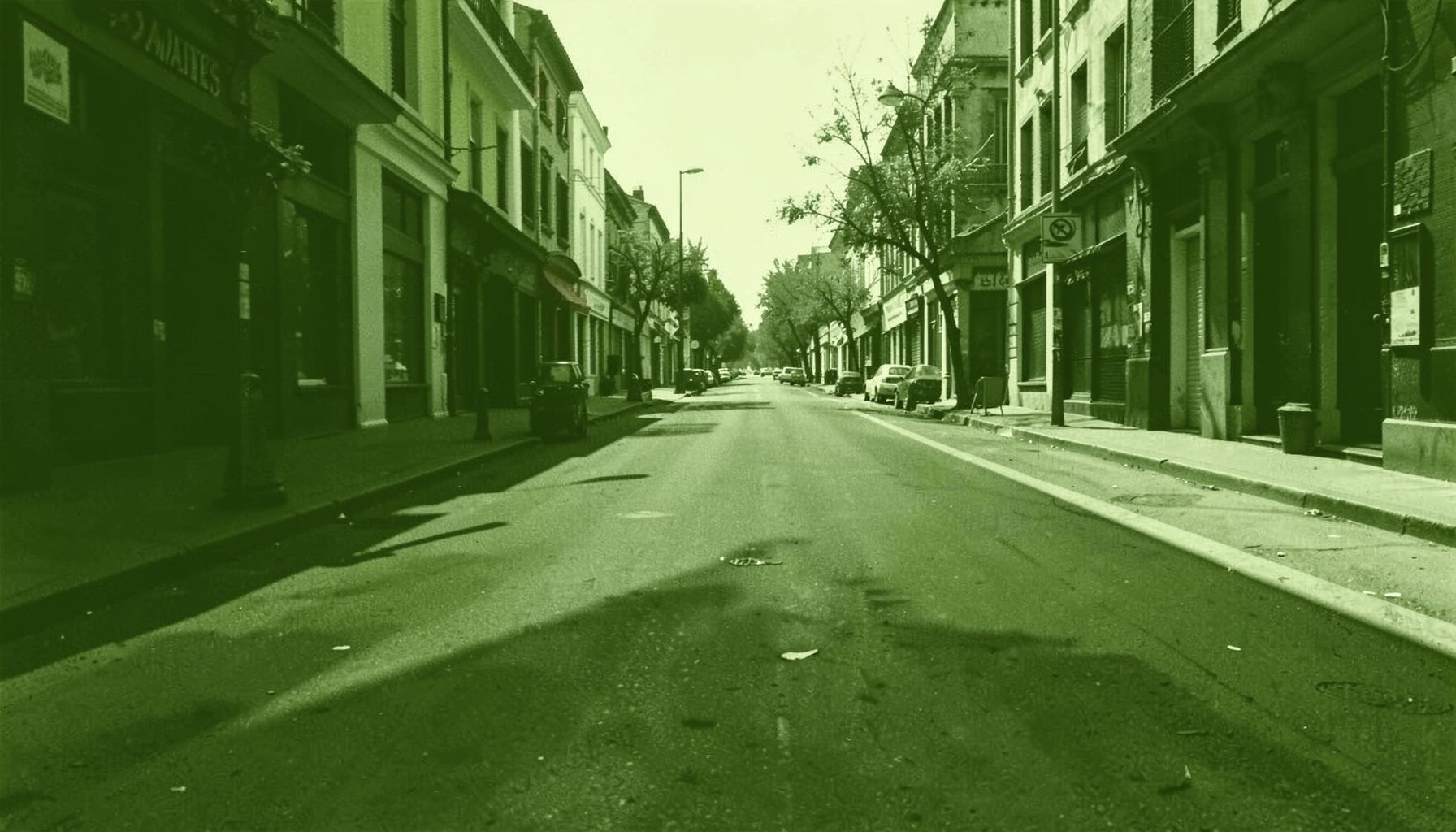 A wide duotone photograph in deep olive green and cream tones showing a calm empty street scene with long shadows, conveying a quiet working atmosphere